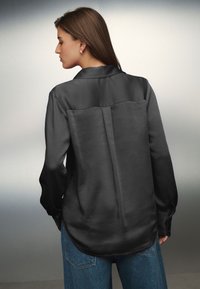 Schwarzes Satinhemd mit langen Ärmeln, Kragen und abgerundetem Saum. Glatte Textur mit einer mittigen Naht am Rücken, kombiniert mit blauen Denim-Jeans.