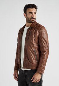 Man met donker haar en een baard, gekleed in een bruine leren jas, witte shirt en zwarte jeans, kijkt naar de zijkant tegen een effen achtergrond.