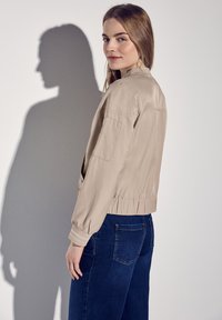 Beige bomber bunda se staženým pasem, dlouhými rukávy a bočními kapsami, v kombinaci s tmavě modrými džíny, které mají hladkou texturu a jemný lesk.