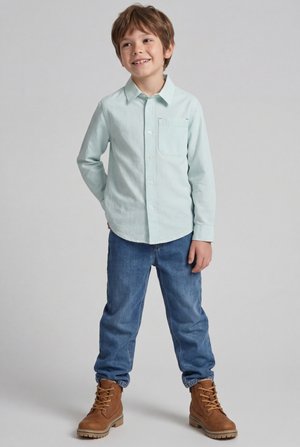 Ragazzo che indossa una maglietta a maniche lunghe verde chiaro a righe, jeans blu e stivali marroni con lacci, in piedi e sorridente su uno sfondo semplice.