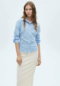 Camicia azzurro chiaro a maniche lunghe con righe verticali, abbinata a una gonna aderente color crema, su uno sfondo neutro.