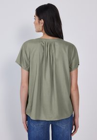 Blusa verde oliva con maniche corte, caratterizzata da un design arricciato sul retro e una texture liscia. Abbinata a jeans blu, mostrata di spalle.