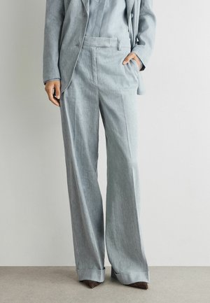 Femme portant un blazer gris clair sur mesure et un pantalon large avec des chaussures marron pointues, debout contre un mur clair uni.