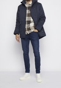 Marinblå vadderad jacka med huva, grå och brun rutad skjorta, mörka jeans och vita högstövlar. Tydliga, funktionella designelement.