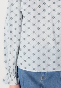 Blusa azul clara com um padrão floral em azul escuro, apresentando mangas bufantes e punhos franzidos. Combinada com jeans de ganga.
