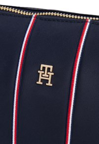 Tamsiai mėlynas poliesterio audinio krepšelis su auksine užtrauktuku, puoštas auksiniu logotipu ir šone esančiomis raudonai, balta ir mėlyna dryžuotomis detalėmis.