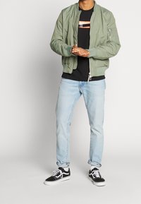 Una chaqueta bomber verde, camiseta negra con logo rojo, jeans azul claro y zapatillas negras con suelas blancas. Atuendo casual, diseño simple.