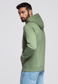 Sudadera verde de tejido suave con un bolsillo canguro, capucha ajustable y puños de canalé, combinada con jeans de color azul claro.