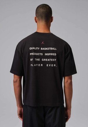 Uomo di spalle che indossa una maglietta nera con testo bianco: "Prodotti da basket di qualità ispirati dal più grande giocatore di sempre" e un logo rosso sopra.