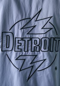 Tessuto grigio con un audace ricamo nero. La parola "DETROIT" è in evidenza, circondata da forme stilizzate e astratte di fulmini.