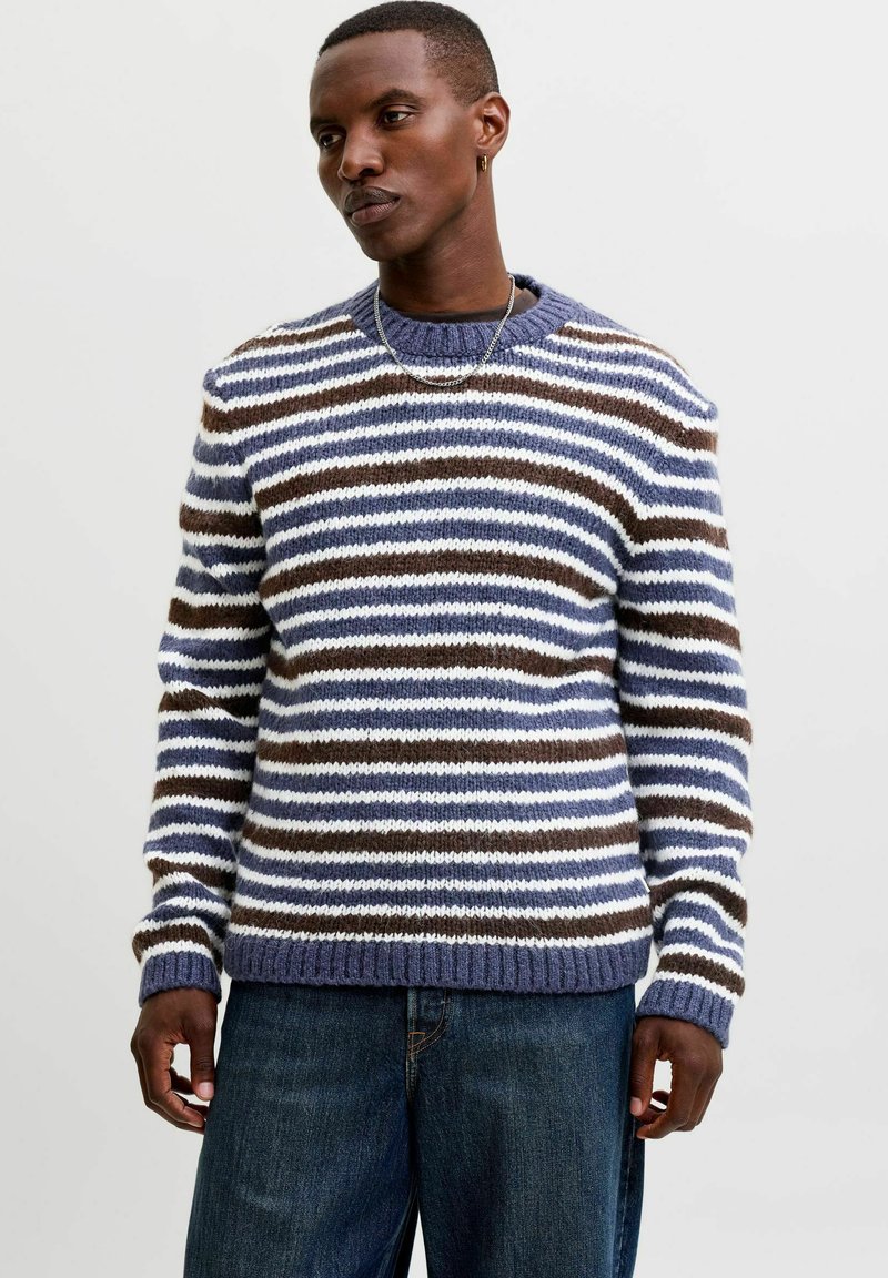 Homme portant un pull en tricot rayé avec des bandes horizontales bleues, blanches et marron et un jean bleu foncé sur un fond uni.