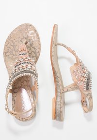 Beżowe płaskie sandały z kwiecistymi wzorami, ozdobione różowymi i srebrnymi koralikami oraz kamieniami, z paskami na kostce i pętlami na palce, pokazane z góry i z boku.