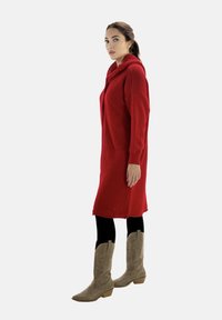 Vestido rojo de punto con capucha y corte holgado, combinado con leggings negros y botas vaqueras de color marrón claro con patrones perforados.