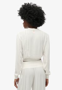 Witte crop top met lange mouwen, gemaakt van een textuurstof, met kantdetails langs de naden en een klein label achter in de hals.