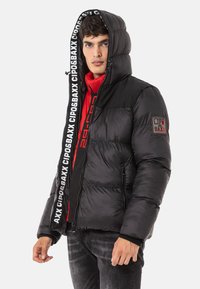 Schwarze Pufferjacke mit rotem Innenfutter und Logo-Details. Mit Kapuze, Reißverschluss und elastischen Bündchen; glatte und glänzende Textur.