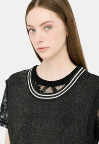 Juodas ir metalizuotas mezginys su nėriniuotomis rankovėmis, su balta juostele ant apykaklės ir sudėtingu nėriniu detalėmis ant iškirptės.