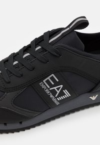 Svart sportig sko med en mix av släta och texturerade material, tydlig logotyp, snörning och randig detalj vid kragen.