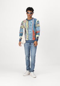 Kleurrijke sweatshirt met blauw, rood en groene patronen. Spijkerbroek en witte sneakers maken de outfit compleet. Lichte tote bag in de hand.