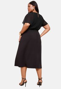 Schwarzes Kleid mit kurzen, durchsichtigen Ärmeln, betonter Taille und knielangem Rock. Verfügt über einen Schlüssellochausschnitt auf der Rückseite und eine glatte Textur. Passt gut zu schwarzen Absätzen.