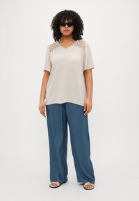 Beige V-halsbluse med blonderdetaljer på skuldrene, parret med løse blå bukser. Modellen bærer sorte solbriller og hvide sandaler.