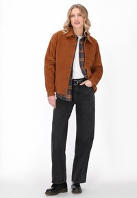 Jonge vrouw in een bruine jas over een geruite shirt en witte top, zwarte wijde jeans, zwarte laarzen, met een hand in haar zak, staand op een witte achtergrond.