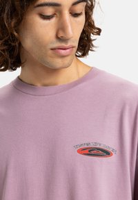 Lilla bomulds t-shirt med et frontgrafik, der viser teksten "Smager som himlen" i sort og rød, rund hals og korte ærmer.