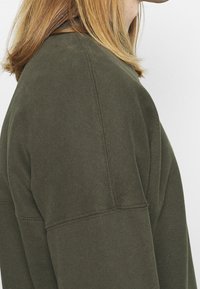 Sudadera verde oliva con cuello redondo, confeccionada en un tejido texturizado, con costuras visibles y un ajuste holgado, mostrada desde un ángulo lateral.