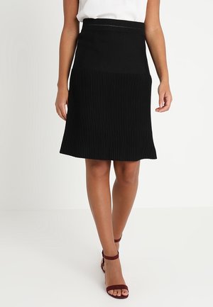 Vrouw draagt een zwarte geplooide knielange rok, witte top en bordeauxrode sandalen met hoge hakken, staand tegen een effen witte achtergrond.