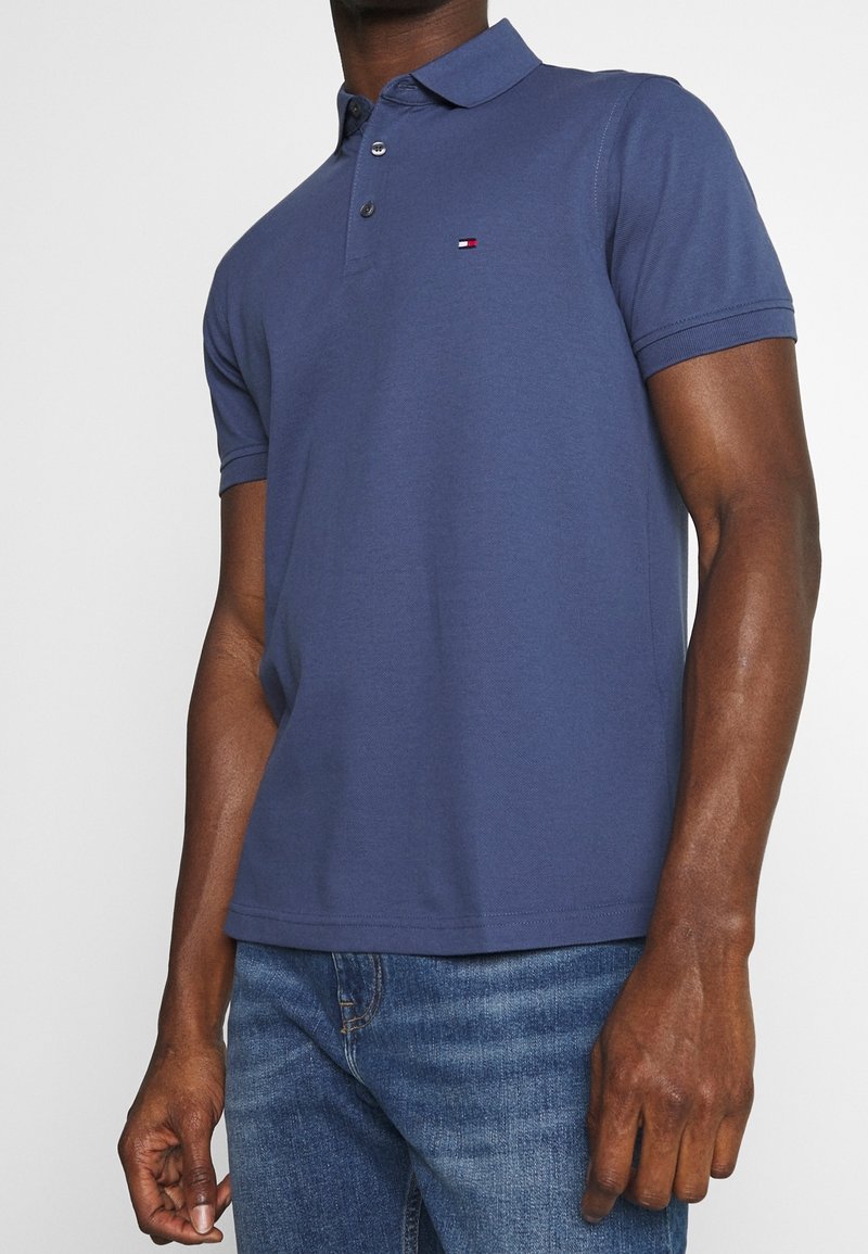 Homme portant un polo bleu marine à manches courtes avec un petit logo de drapeau sur la poitrine et un jean en denim bleu.