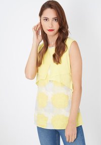 Top sin mangas de color amarillo con diseño en capas, que presenta una tela estampada más clara debajo con motivos florales en azul pálido y amarillo.
