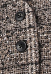 Brūns un melns tveida audums ar nedaudz pūkainu tekstūru, kas rotāts ar divām melnām četrcauruļu pogām.