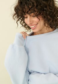 Helles blaues Sweatshirt mit entspannter Passform, gerippten Bündchen und sichtbaren Nähten. Der Stoff wirkt weich mit einer strukturierten Oberfläche.