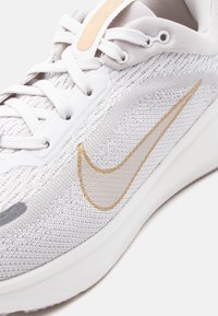 Κοντινό πλάνο ενός λευκού και μπεζ παπουτσιού Nike για τρέξιμο με ύφασμα από δίχτυ, λευκά κορδόνια και μπεζ λογότυπο Nike στο πλάι.