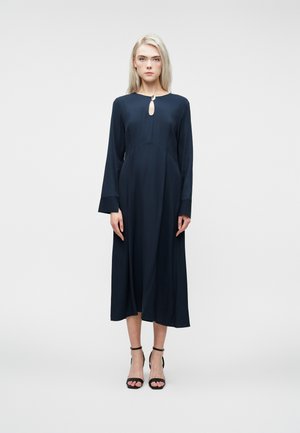Woman wearing a long-sleeve, navy midi dress with a front keyhole detail fastened by a small silver clasp, and black ankle-strap heels.