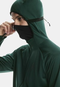 Mann, der eine schwarze Gesichtsmaske unter einer dunkelgrünen Kapuzenjacke mit Reißverschluss anpasst, blickt nach links vor einem weißen Hintergrund.