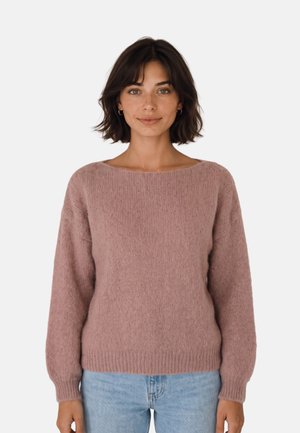 Mujer con cabello corto y oscuro, vistiendo un suéter de punto color malva y vaqueros azul claro, de pie frente a un fondo blanco liso.