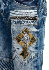 Dunkelblaue Jeans mit einem hellblauen Wascheffekt, die ein auffälliges besticktes Kreuzdesign in Gold und Schwarz sowie einen Reißverschlusstaschen-Detail aufweisen.