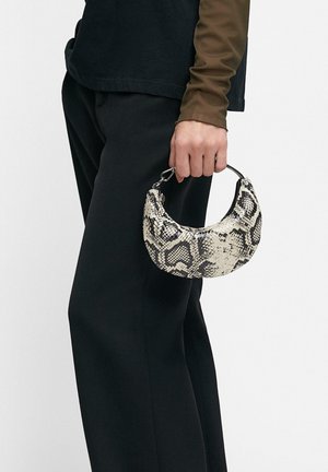 Osoba ubrana w czarne spodnie i brązową bluzkę z długim rękawem trzyma małą, półksiężycową torbę z wzorem czarno-białej skóry węża.