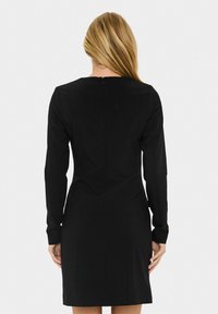 Femme aux longs cheveux blonds portant une robe noire ajustée à manches longues, vue de dos contre un fond blanc uni.