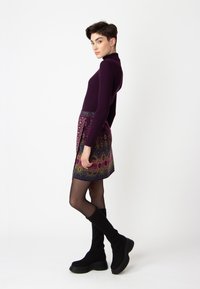 Lila gerippter Rollkragenpullover mit einem bunt gemusterten Rock, der aufwendige Designs und einen schwarzen Taillenbund aufweist, kombiniert mit knielangen schwarzen Stiefeln.