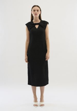 Black midi dress with textured fabric, cap sleeves, and a keyhole detail at the neckline. Styled with white open-toe shoes.