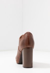 Scarpa con tacco in pelle marrone dalla forma arrotondata, tacco in legno a righe verticali e cuciture dettagliate sul retro.