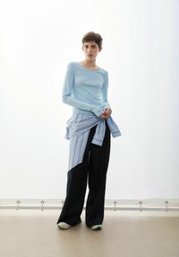 Hellblaues Langarmshirt kombiniert mit einem gestreiften Hemd, das in der Taille gebunden ist. Weite, dunkel gestreifte Hose, grünlich getönte Schuhe vervollständigen das Outfit.