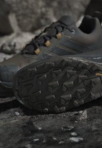 Gros plan d’une chaussure de randonnée noire montrant une semelle crantée robuste et une partie de la tige avec des lacets sur un sol rocheux.