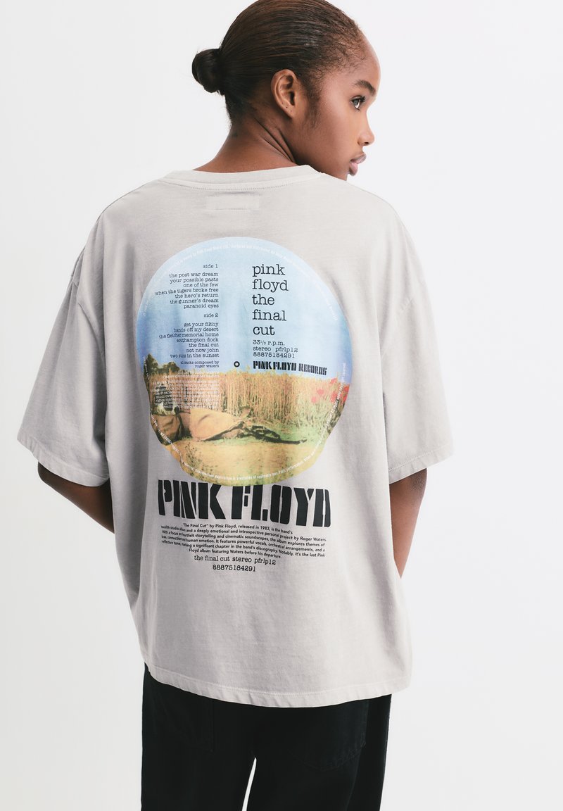 Mladić s kosom u punđi nosi bež majicu s grafikom Pink Floyd "The Final Cut", blago okrenut udesno protiv jednostavne pozadine.