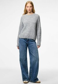 Jauna sieviete, ģērbusies pelēkā adītā džemperī, platajās zilos džinsos un bēšās sporta kurpēs, stāv uz balta fona.