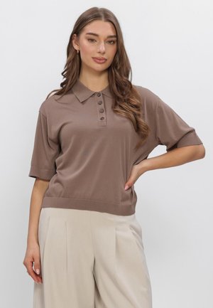 Kobieta o długich brązowych włosach, ubrana w brązową polo z krótkim rękawem i guzikami oraz beżowe plisowane szerokie spodnie, pozująca z ręką na biodrze.