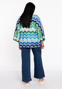 Bunt gestreifter Kimono mit grün, blau und weißen Tönen, kombiniert mit dunkelblauen weit geschnittenen Jeans und hellen Slippern.