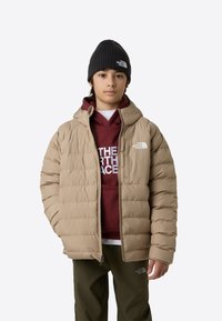 Geacă matlasată bej cu fermoar frontal, glugă și manșete striate, purtată peste un hanorac burgund. Modelul poartă o căciulă neagră și pantaloni verde-olive.