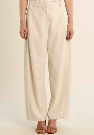 Pantaloni beige a gamba larga con pieghe frontali indossati da una persona in piedi, abbinati a sandali tan con punta aperta.